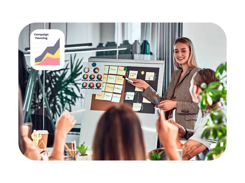 A young professional leads a collaborative campaign planning session using a whiteboard and sticky notes, while colleagues engage enthusiastically, demonstrating teamwork and creativity in apprenticeships.