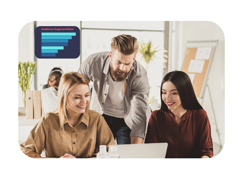 Three young professionals collaborating around a laptop in a bright, modern office setting, with a chart labeled "Audience Segmentation" on the wall, illustrating teamwork and learning.