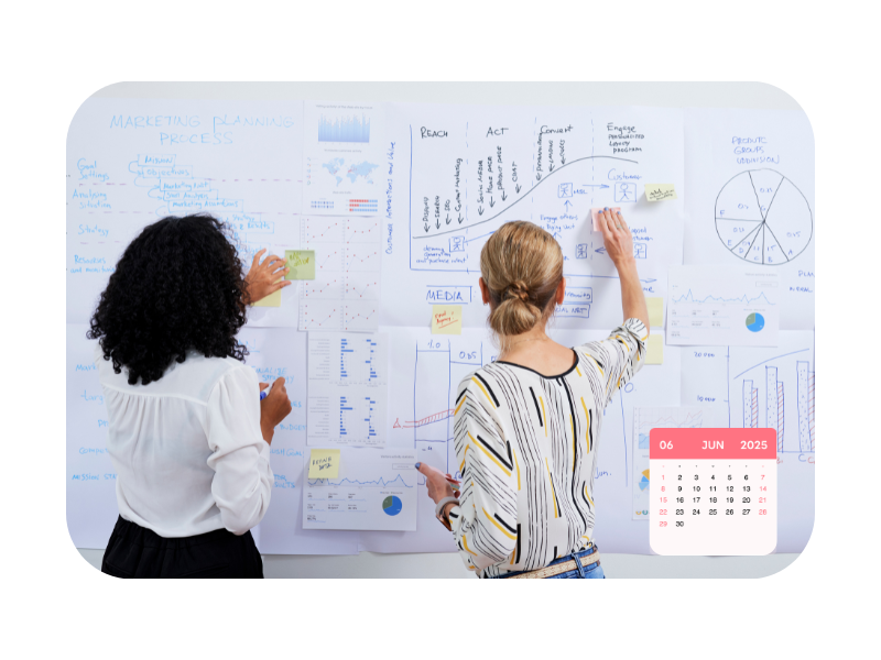 Two women planning marketing strategies on a whiteboard filled with charts, graphs, and notes, representing practical training in a multi-channel marketer apprenticeship.